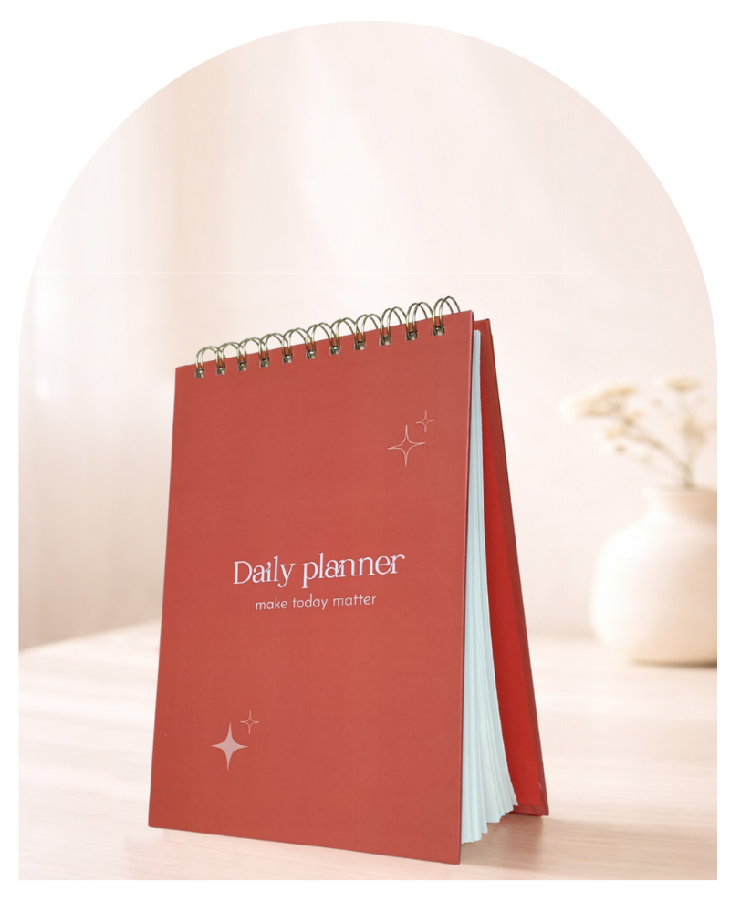 A red spiral-bound daily planner standing upright on a light wooden surface, with white text reading 'Daily planner' and 'make today matter' on the cover. The background is softly blurred with a neutral beige tone and a small vase.
