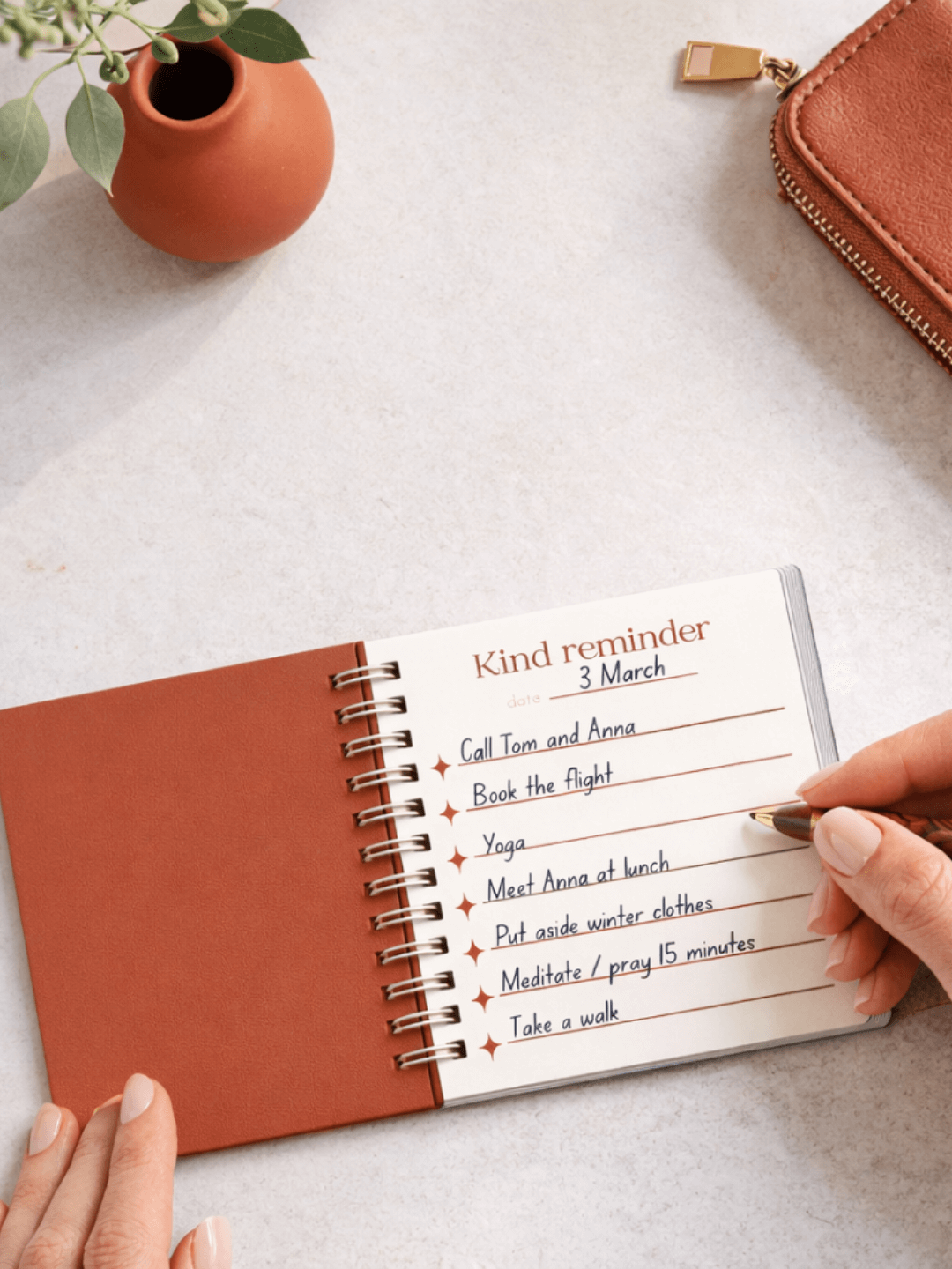 Open spiral-bound notebook with a kind reminder list on a light surface, with a small vase and brown pouch in the background.